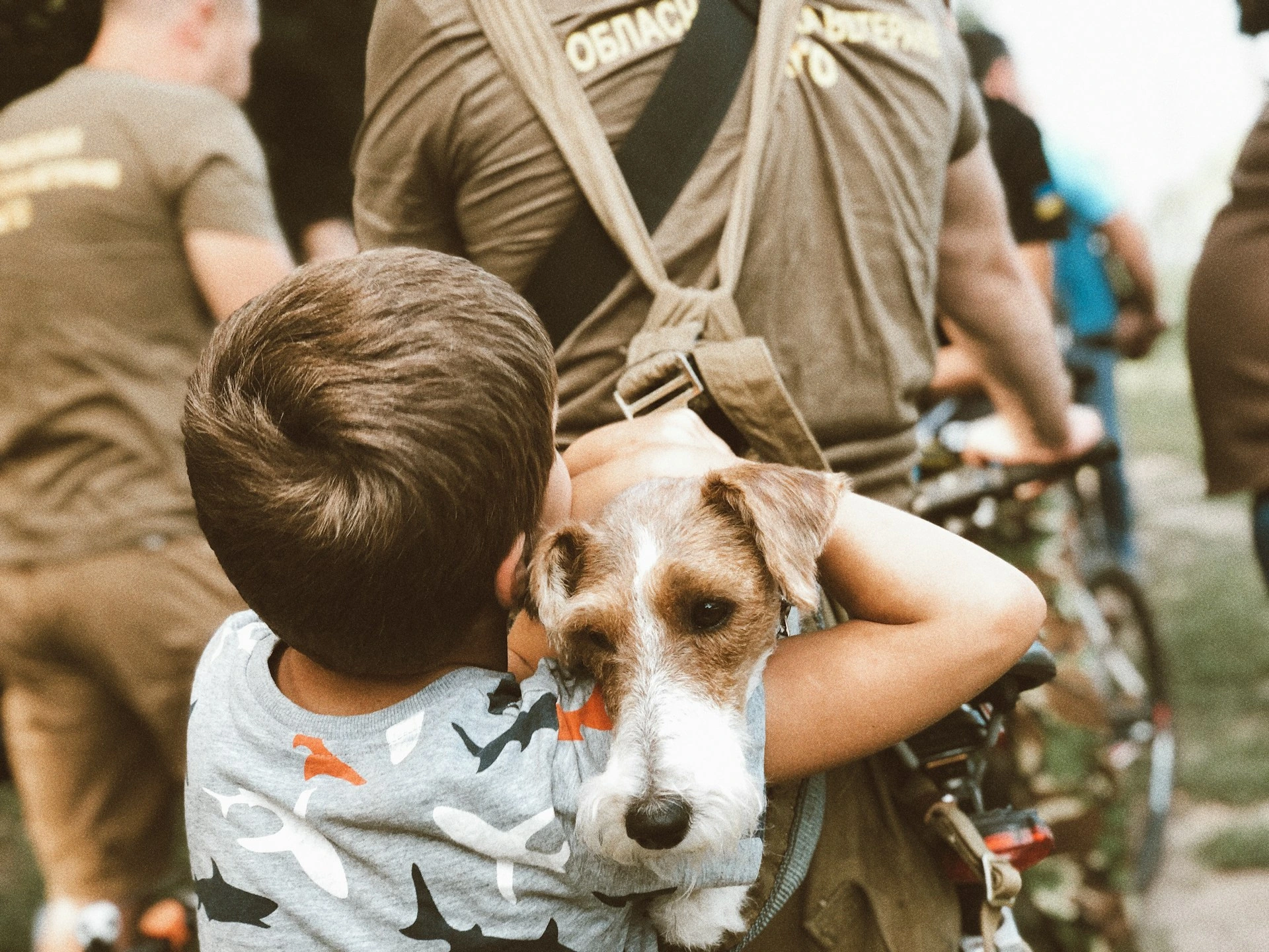 A child in a shark T-shirt hugs a scruffy, white and brown dog that is held in a person's arms.